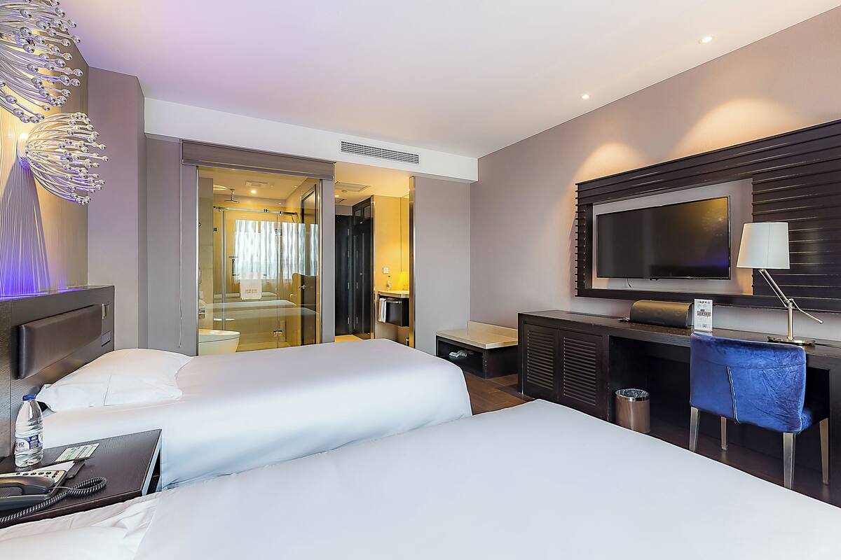 A hotel room with two beds, a TV, a desk, and a bathroom visible through a glass door.