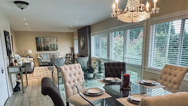 A dining area with a glass table, cushioned chairs, a chandelier, and large windows with blinds. - 3BR Edgemont Retreat | Views, Deck, Near Grouse (North Vancouver)