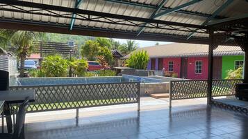 A covered outdoor area with a tiled floor, a pool, and a view of colorful buildings and greenery.