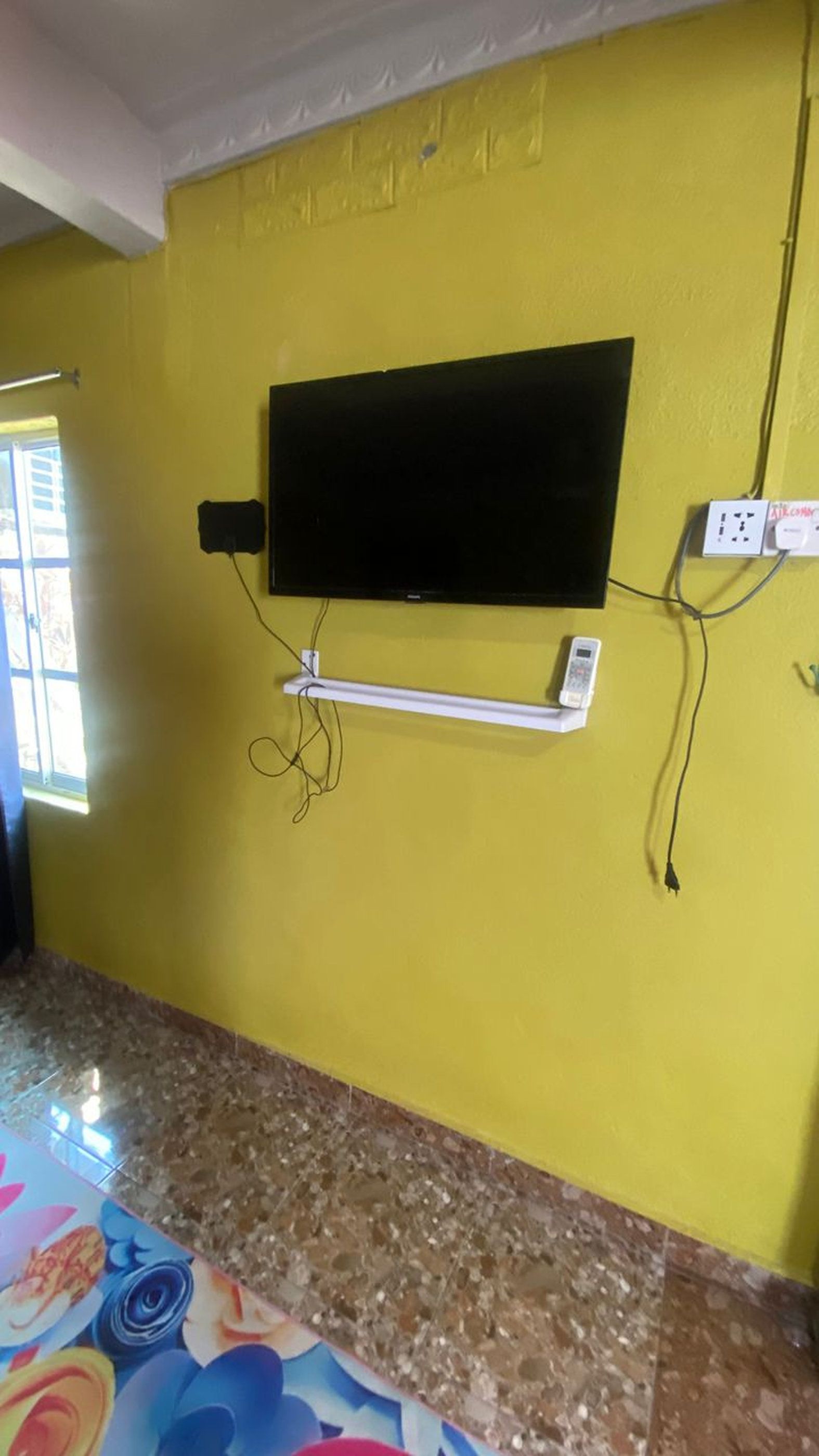 A room with a yellow wall, a flat-screen TV mounted on it, a power outlet, and a granite countertop.
