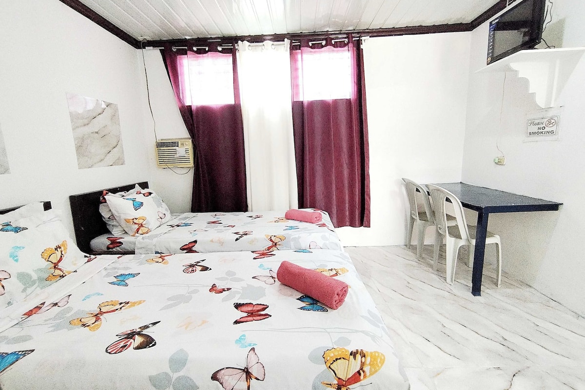 A bedroom with a bed, a desk, and a chair. There are curtains on the windows, and a sign on the wall.