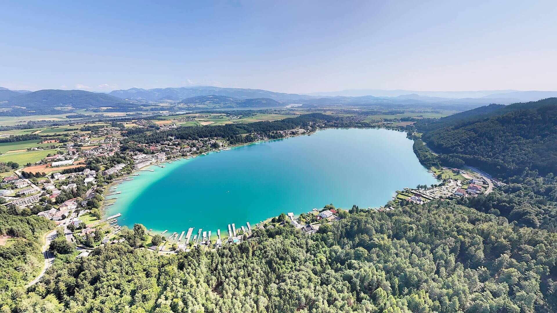 Une vue aérienne d’un lac entouré de collines boisées, avec une petite ville et des bateaux amarrés le long de la rive.