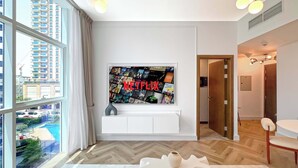 Una sala moderna con un televisor grande mostrando un mural de Netflix, un piso de madera en espiga y vistas a una zona residencial a través de amplias ventanas.