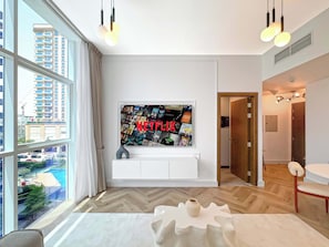 Ein modernes Wohnzimmer mit einem großen Fernseher, der eine Netflix-Wandkunst zeigt, einem Holzboden im Fischgrätmuster und Blick auf eine Wohngegend durch große Fenster.