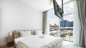Un dormitorio moderno con una cama grande, una mesita de noche, un televisor en la pared y vistas de la ciudad a través de amplias ventanas.