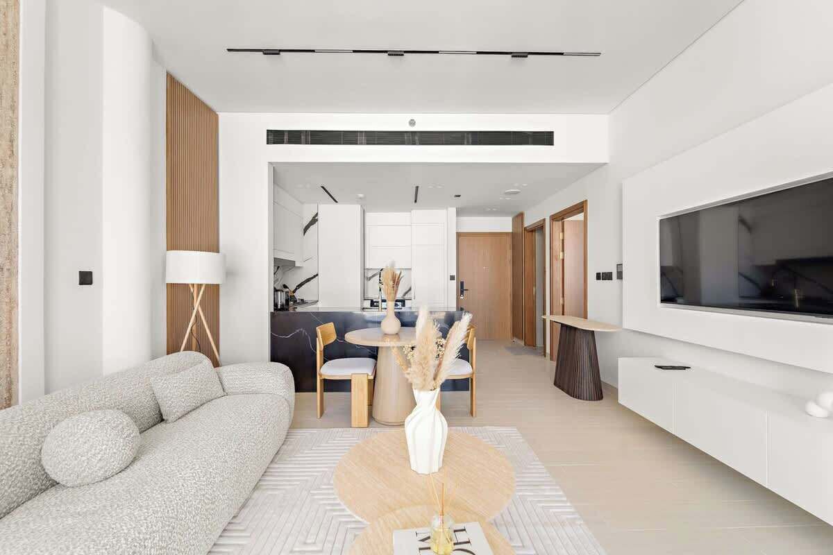 Una sala moderna con un sofá, una mesa de comedor con sillas, un televisor montado en la pared y una cocina al fondo.