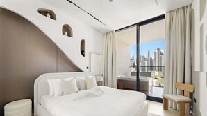 Una habitación de hotel moderna con una cama grande, un banco, una mesita y vistas a la ciudad.