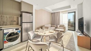 A modern kitchen with a built-in washer, a dining area with round table and chairs, a flat-screen TV, and a large window with curtains. - Modern 1BR in Brand-New Cloud Tower in JVT (Dubai)
