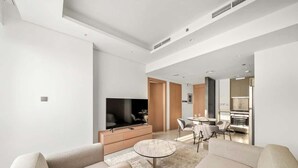 A modern living room with a sofa, a TV, a dining area with chairs, and a kitchen in the background. - Modern 1BR in Brand-New Cloud Tower in JVT (Dubai)