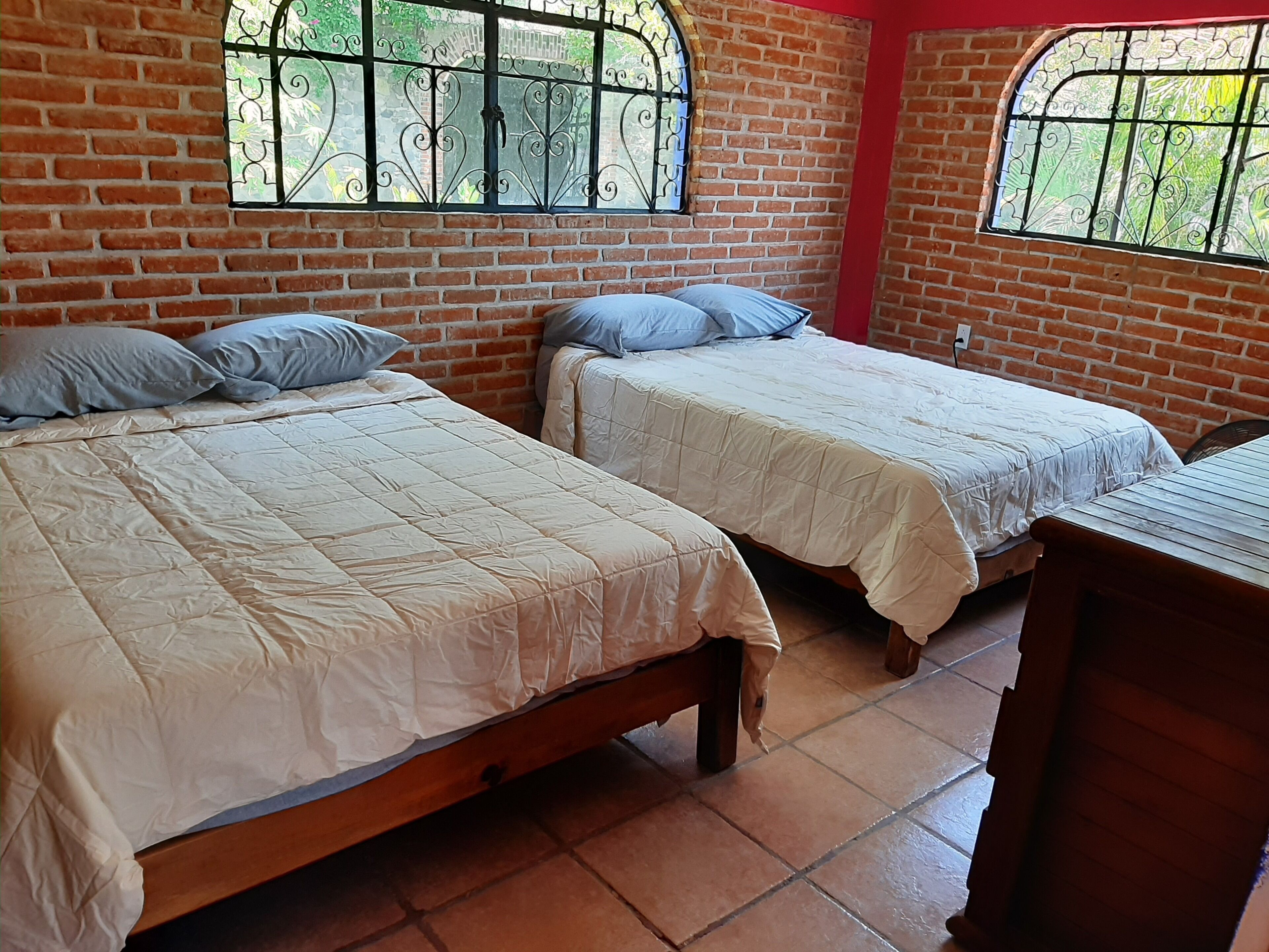 A room with two beds, a wooden desk, and a brick wall with windows.