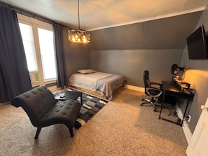 A bedroom with a bed, a desk with a chair, a black armchair, a patterned rug, and a hanging light fixture.