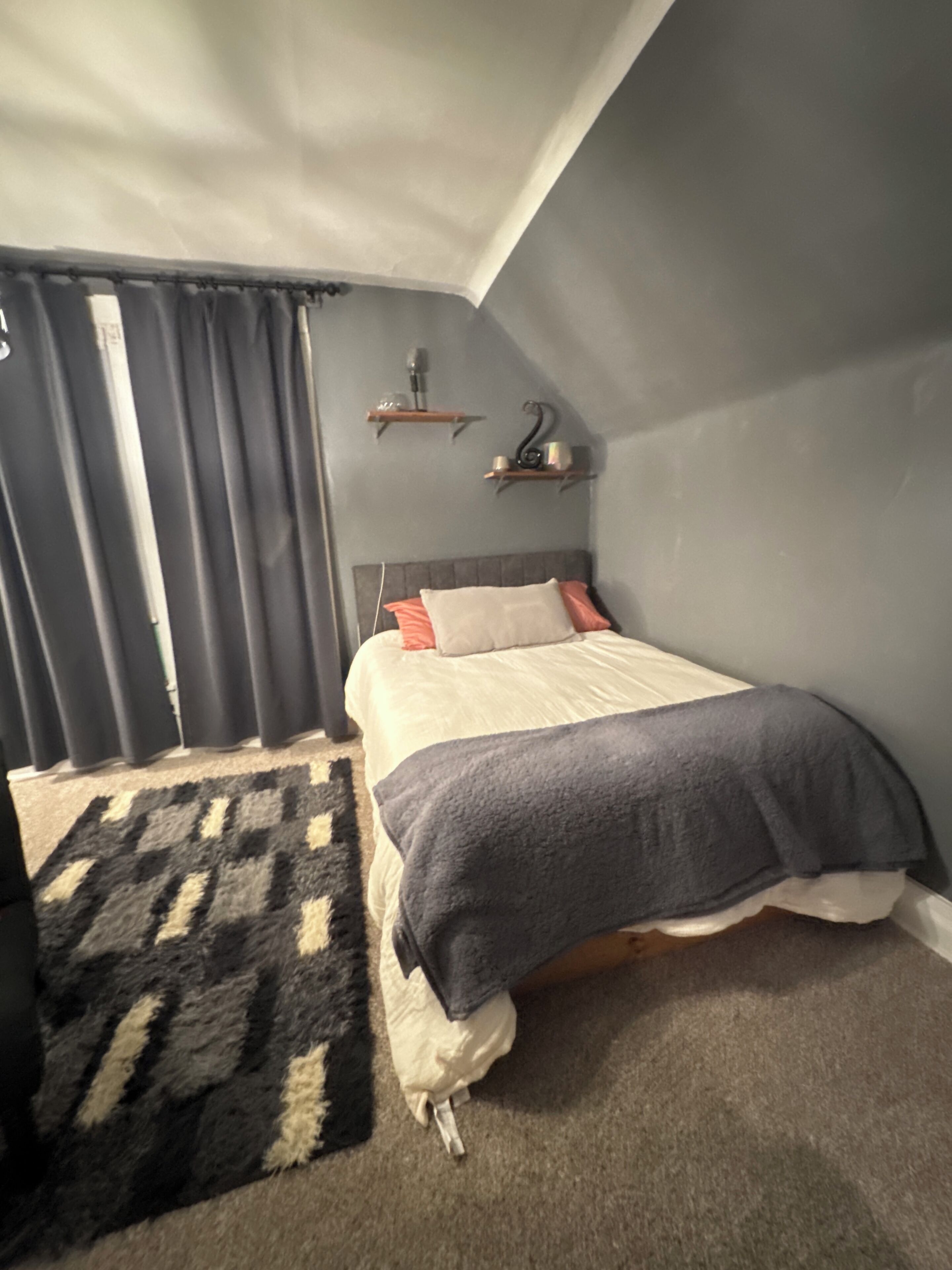 A bedroom with a bed, a rug, a window with curtains, and a wall-mounted shelf.
