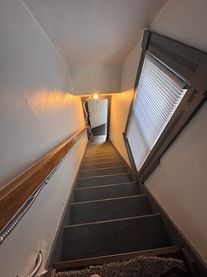 Una escalera estrecha con barandilla, una puerta arriba y una ventana con persianas.