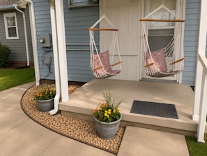 A porch with two hanging chairs, a concrete step, a doormat, and potted plants.