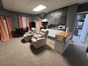 A living room with a sofa set, a dining table with chairs, a kitchen area with cabinets and appliances, and a small wooden table.
