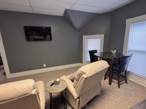 A living room with a gray wall, a flat-screen TV mounted above a fireplace, a round table with chairs, and a beige sofa.
