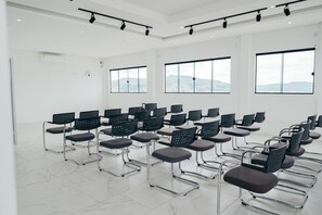 Un salón de conferencias con filas de sillas, ventanas amplias e iluminación en el techo.