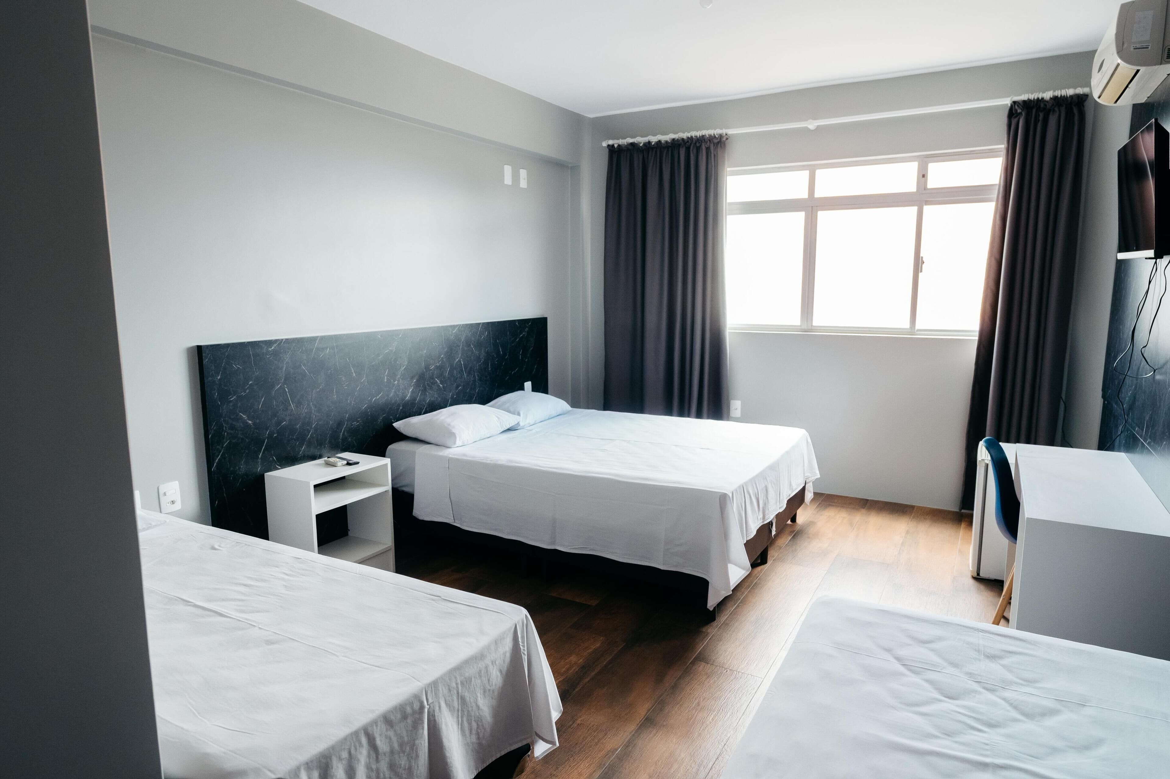 Quarto de hotel com duas camas, uma escrivaninha, uma cadeira e uma janela com cortinas.
