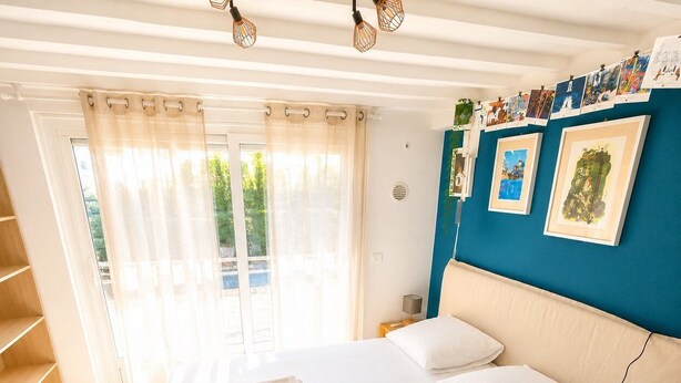 A bedroom with a large bed, a wooden nightstand, a blue accent wall, and a view through the window. - Maison 8p avec jardin, 2 salons & parking (Sainte-Geneviève-des-Bois)