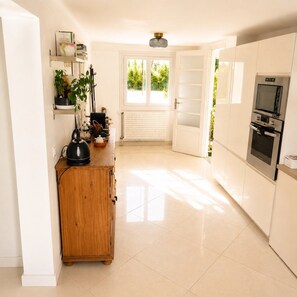 A modern kitchen with white cabinets, a wooden cabinet, a kettle, and a window with blinds. - Maison 8p avec jardin, 2 salons & parking (Sainte-Geneviève-des-Bois)