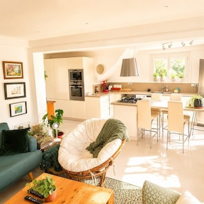 Cozinha moderna com armários brancos, área de jantar com cadeiras e sala de estar com sofá e poltrona verde.