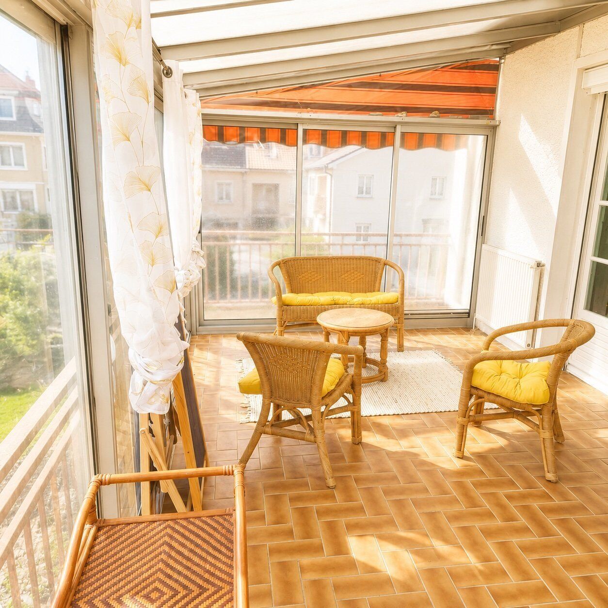 Ein Balkon mit Korbmöbeln, Blick auf Gebäude und ein Fenster mit Vorhang.
