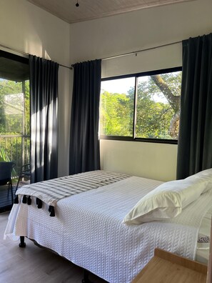 Une chambre à coucher avec un lit, une fenêtre avec des rideaux, une chaise et une vue sur la verdure à l’extérieur.