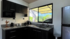 A modern kitchen with dark cabinets, a stainless steel refrigerator, a window with a view of greenery, and a granite countertop. - Tropical Forest Experience with Hot Tub, AC & Terrace (Atenas)