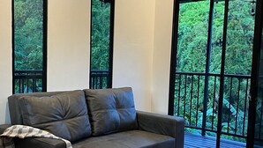 A modern living room with a grey sofa, large windows, and a balcony with a view of greenery. - Tropical Forest Experience with Hot Tub, AC & Terrace (Atenas)