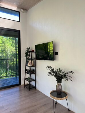 A room with a large window, a bookshelf, a mounted TV, and a small table with a vase. - Tropical Forest Experience in Atenas (Atenas)