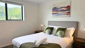Una cama bien hecha con almohadas, una manta y una toalla. Hay un cuadro en la pared, una ventana con vistas al verdor y una mesita de noche con una lámpara.