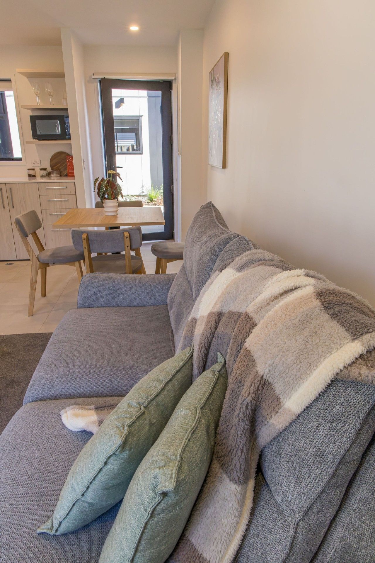 A living room with a sofa, a dining table with chairs, a kitchen area, and a door leading outside.