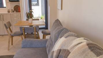 A living room with a sofa, a dining table with chairs, a kitchen area, and a door leading outside.