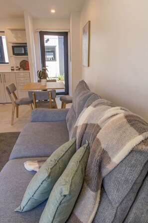 A living room with a sofa, a dining table with chairs, a kitchen area, and a door leading outside. - Stylish Townhouse for Families & Groups (Lower Hutt)
