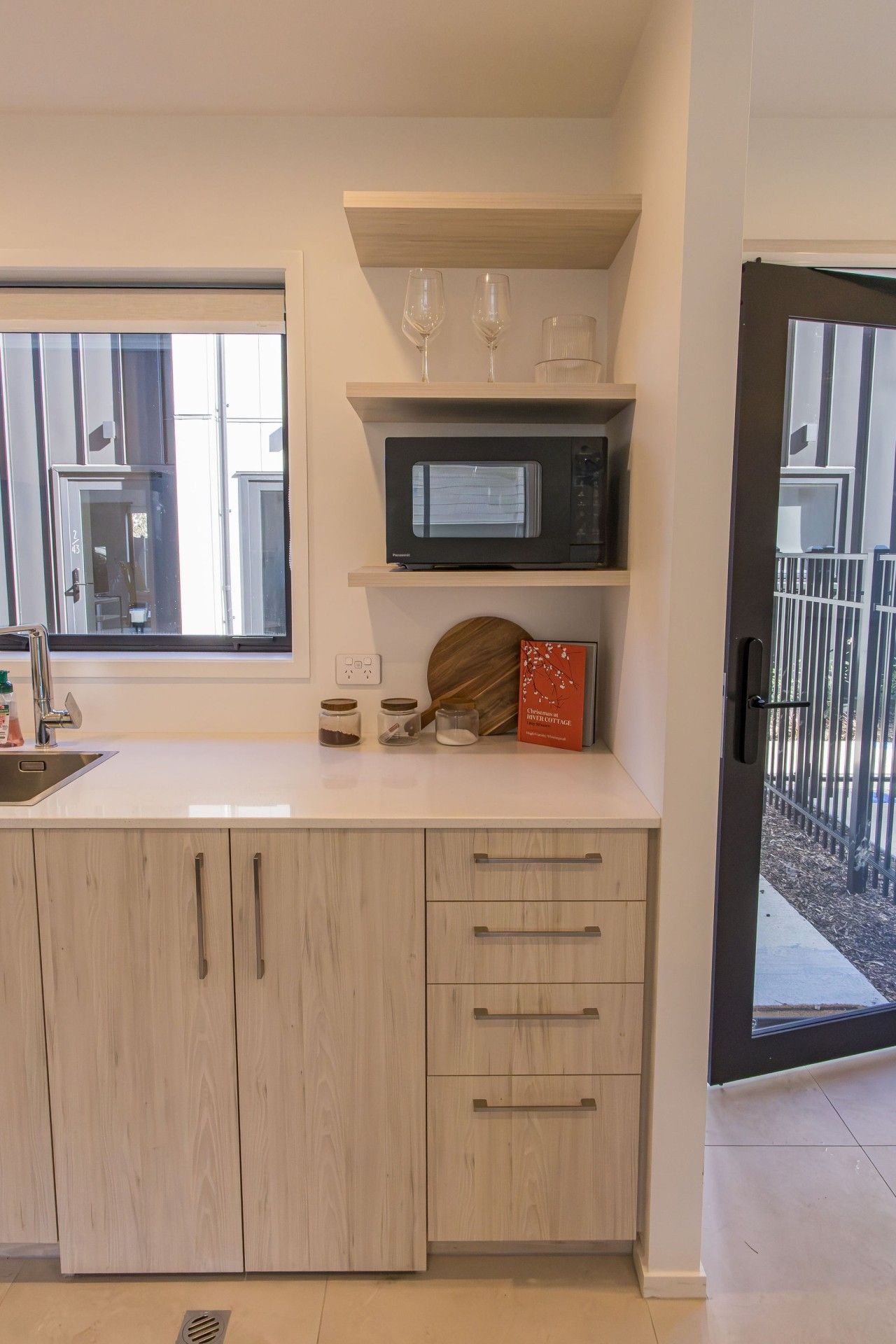 A modern kitchen with a built-in microwave, a sink, and a window. There are jars on the counter and a book titled 'The Art of Living'.