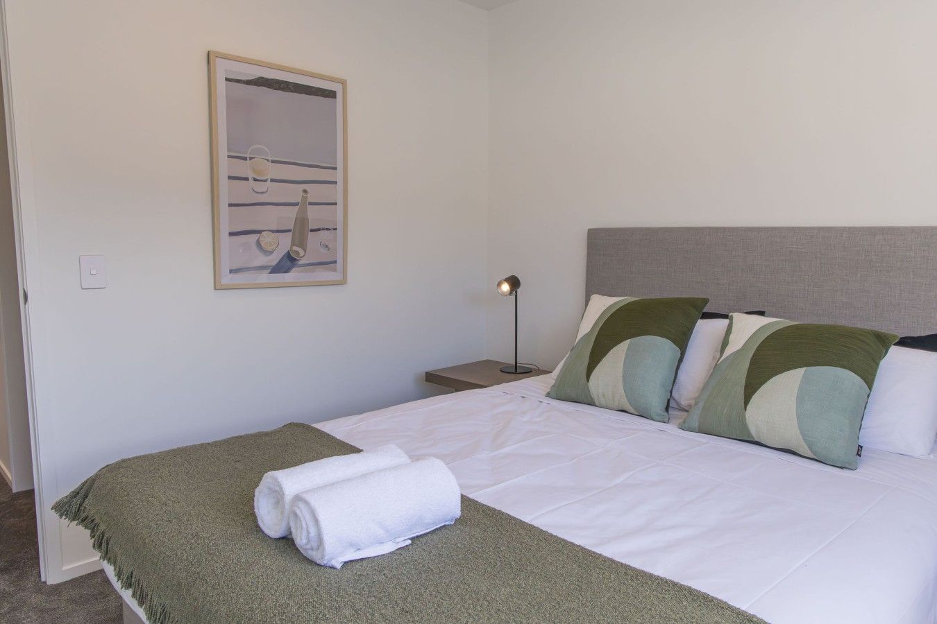 A neatly made bed with a green throw blanket and rolled white towels, a bedside lamp, and a framed picture on the wall.
