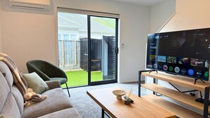 Una sala de estar con un sofá, una mesa de centro, un televisor sobre un mueble bajo y un ventanal corredizo que da a un patio trasero.