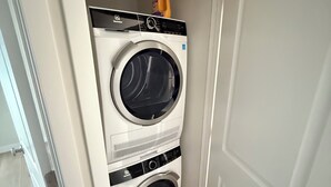 Una lavanderia piccola e funzionale con due lavatrici affiancate, un ripiano con una bottiglia e una porta che dà su un’altra stanza.