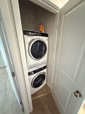 A compact laundry room with two stacked front-loading washing machines, a small shelf with a bottle, and a door leading to another room.