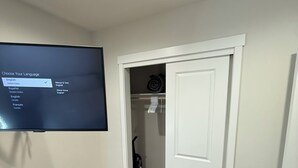 Un corridoio con una televisione che mostra le opzioni di selezione della lingua, un armadio con vestiti e un aspirapolvere, e una porta che conduce in un'altra stanza.