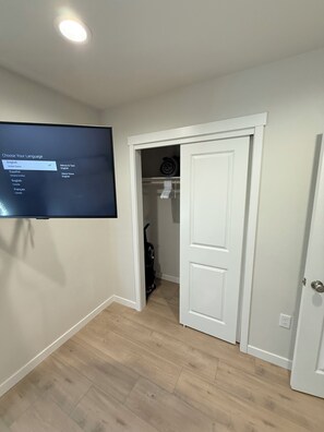 A hallway with a television displaying language selection options, a closet with clothes and a vacuum cleaner, and a door leading to another room.