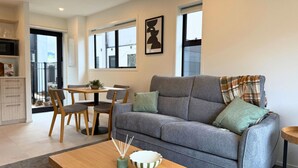 A modern living room with a grey sofa, wooden dining table, and a kitchen area in the background. - Wellington Retreat with Private Courtyard (Lower Hutt)