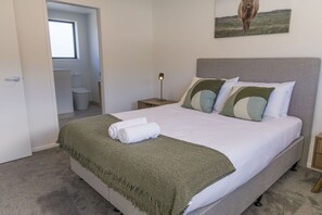 A bedroom with a bed, a nightstand, a bedside lamp, a towel, a blanket, a picture on the wall, and a door leading to a bathroom. - Fairfield Townhouse with Private Courtyard (Lower Hutt)