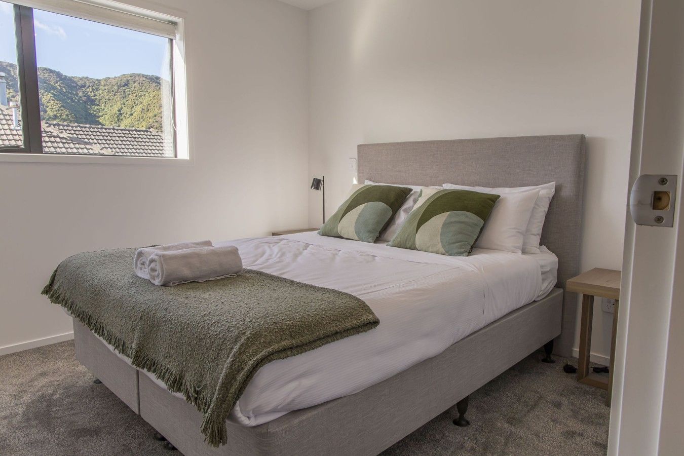A neatly made bed with a green throw blanket, pillows, and a rolled towel.
