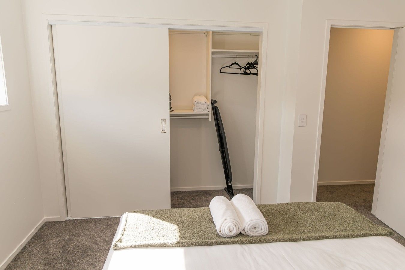 A bedroom with a bed, a green area rug, a closet with a hanging rack, and a folded towel on the bed.