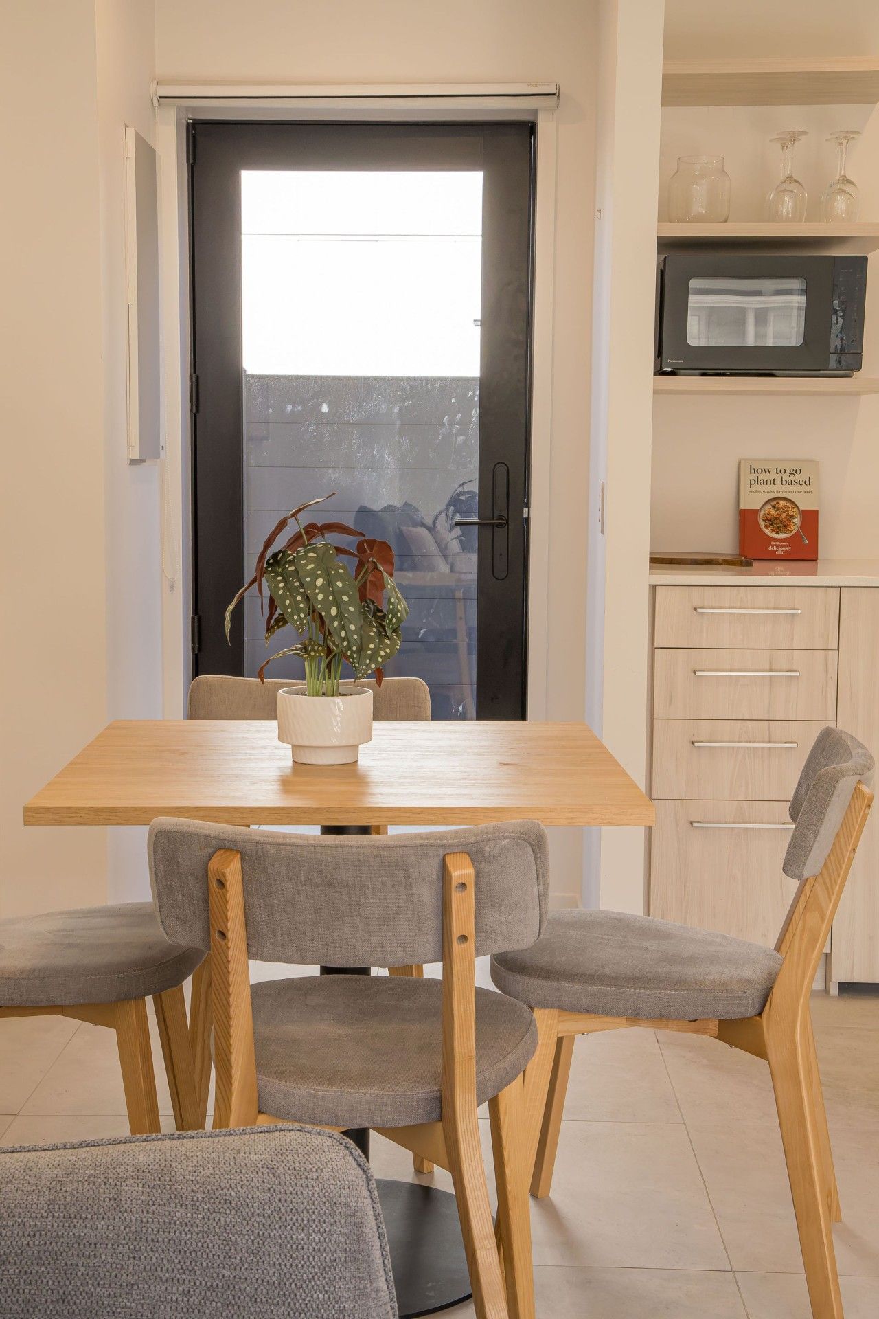 A dining area with a wooden table and four grey upholstered chairs. A potted plant with green leaves and white spots is on the table.