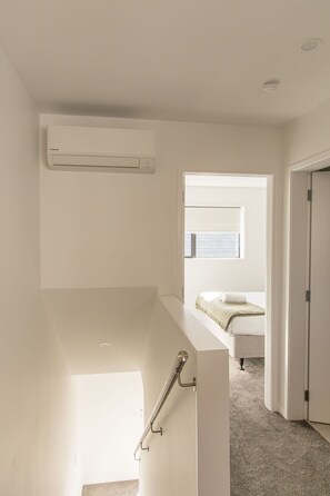 Une chambre moderne et minimaliste, dotée d’un système de climatisation intégré, d’une fenêtre avec stores, d’un lit recouvert d’une couette blanche et d’une porte donnant sur une autre pièce.