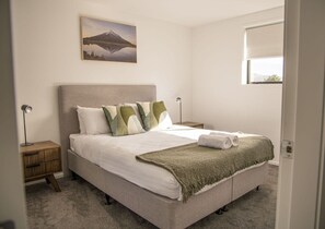 A bedroom with a bed, pillows, a blanket, a nightstand, a lamp, and a painting on the wall. - Modern 2BR Townhouse in Fairfield (Lower Hutt)