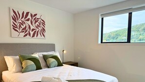 Una cama bien hecha con almohadas, una manta y una toalla enrollada. Se ve una ventana con vistas a un paisaje verde.