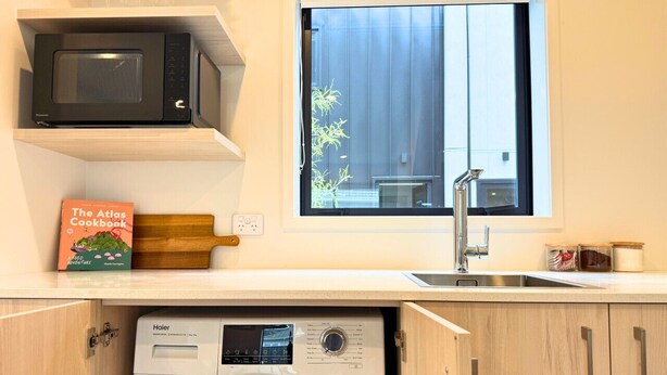 Una cocina compacta con microondas, una lavadora Haier, una ventana con vistas y un libro titulado 'The Atlas Cookbook'.
